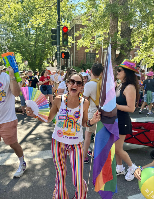 Chelsea at a pride parade, smiling, holding a progress flag and fan, wearing colorful stripes.